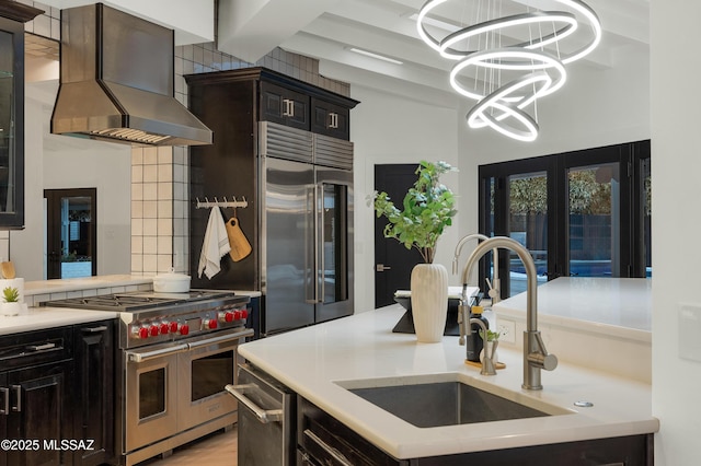 kitchen with wall chimney range hood, an inviting chandelier, decorative backsplash, sink, and high quality appliances