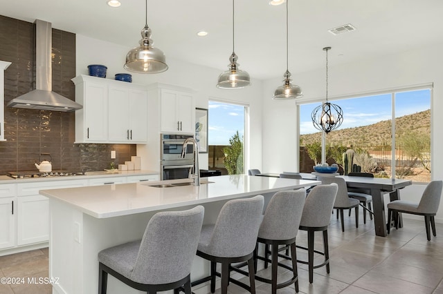 kitchen featuring decorative light fixtures, a center island with sink, appliances with stainless steel finishes, and wall chimney range hood