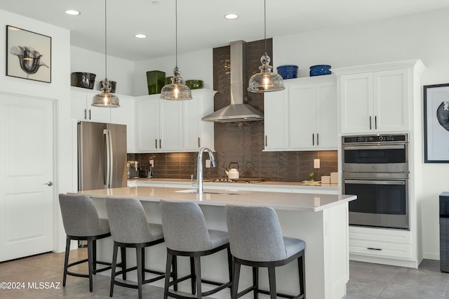 kitchen with stainless steel appliances, white cabinetry, wall chimney exhaust hood, and an island with sink