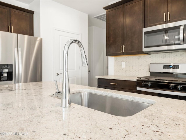 kitchen with decorative backsplash, light stone countertops, stainless steel appliances, dark brown cabinets, and a sink