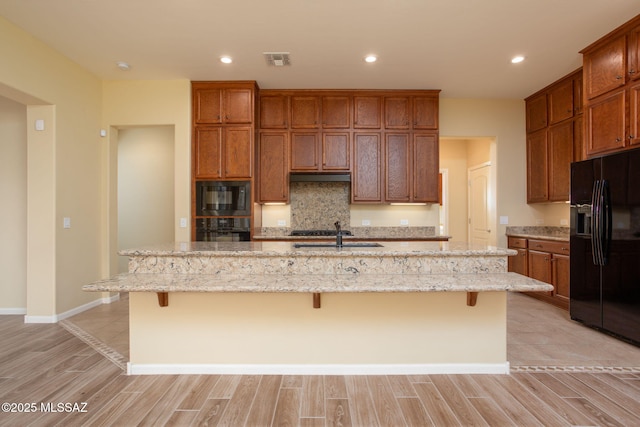 kitchen featuring light stone counters, a breakfast bar, an island with sink, and black appliances