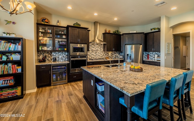 kitchen featuring stainless steel appliances, beverage cooler, a kitchen island with sink, and wall chimney range hood