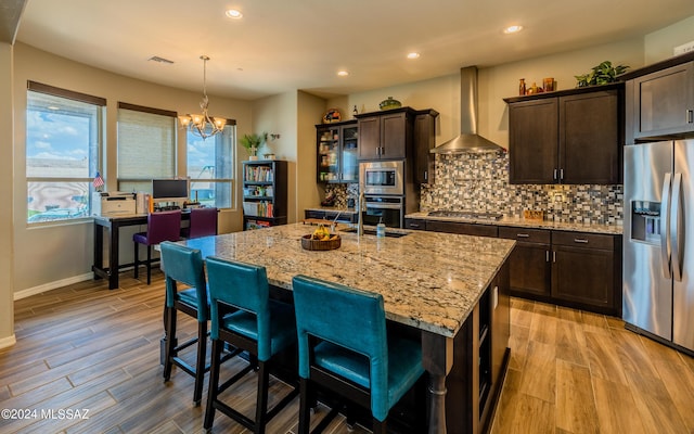 kitchen featuring decorative light fixtures, wall chimney range hood, stainless steel appliances, light stone countertops, and a center island with sink