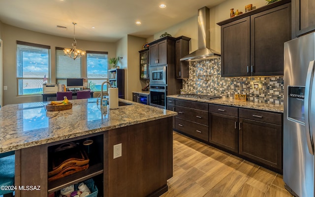 kitchen featuring light stone counters, wall chimney exhaust hood, dark brown cabinetry, stainless steel appliances, and a kitchen island with sink