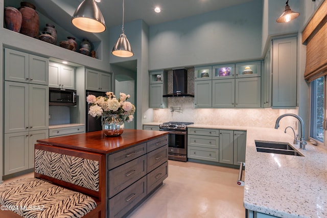 kitchen with light stone counters, wall chimney exhaust hood, stainless steel appliances, sink, and decorative light fixtures