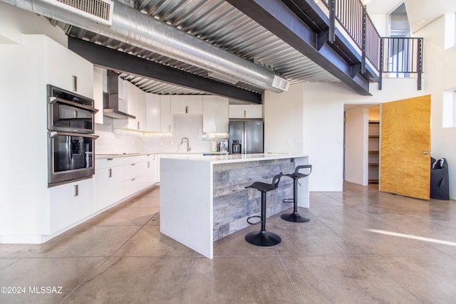 kitchen with white cabinetry, wall chimney exhaust hood, a breakfast bar, stainless steel refrigerator with ice dispenser, and multiple ovens