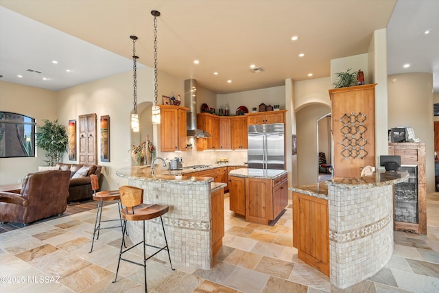 kitchen with built in fridge, wall chimney range hood, kitchen peninsula, pendant lighting, and a breakfast bar