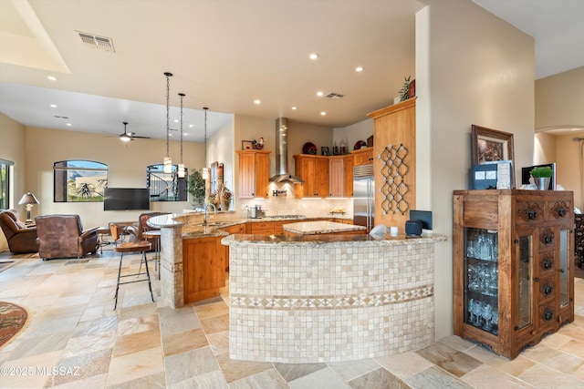 kitchen featuring light stone countertops, appliances with stainless steel finishes, wall chimney range hood, hanging light fixtures, and kitchen peninsula