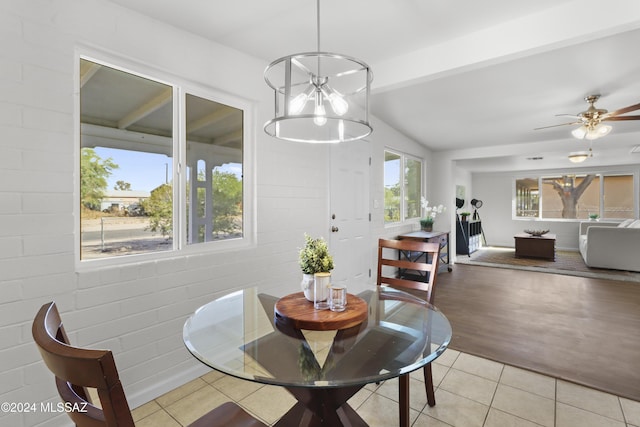 tiled dining space with vaulted ceiling with beams, brick wall, a healthy amount of sunlight, and ceiling fan with notable chandelier