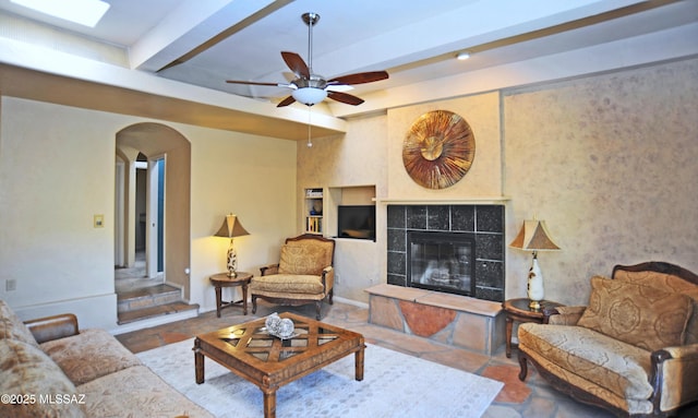 living room with a tiled fireplace, ceiling fan, and beamed ceiling