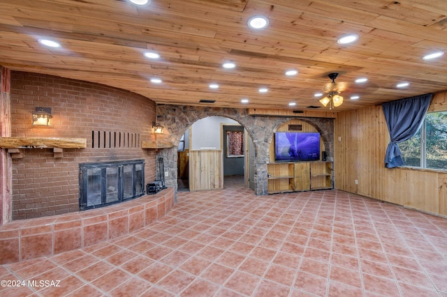 unfurnished living room with a large fireplace, wooden ceiling, ceiling fan, and wooden walls