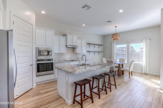 kitchen with white cabinetry, stainless steel appliances, light stone counters, decorative light fixtures, and a center island with sink