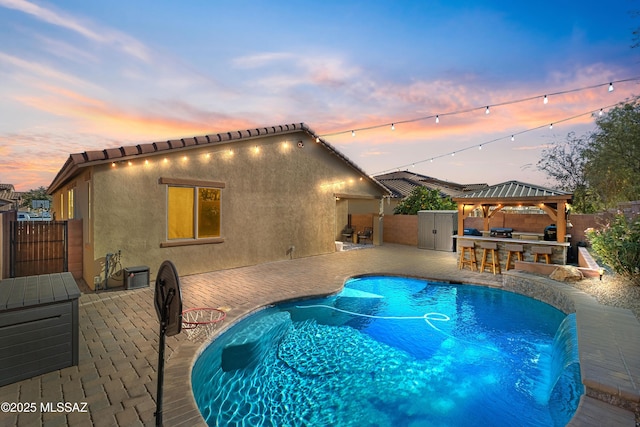 pool at dusk featuring a gazebo, a bar, a patio, and pool water feature