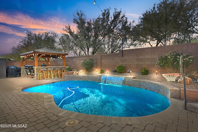 pool at dusk with area for grilling, pool water feature, a gazebo, a patio, and an outdoor bar