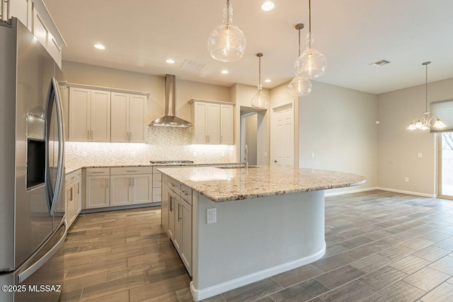 kitchen with appliances with stainless steel finishes, tasteful backsplash, a kitchen island with sink, hanging light fixtures, and wall chimney range hood