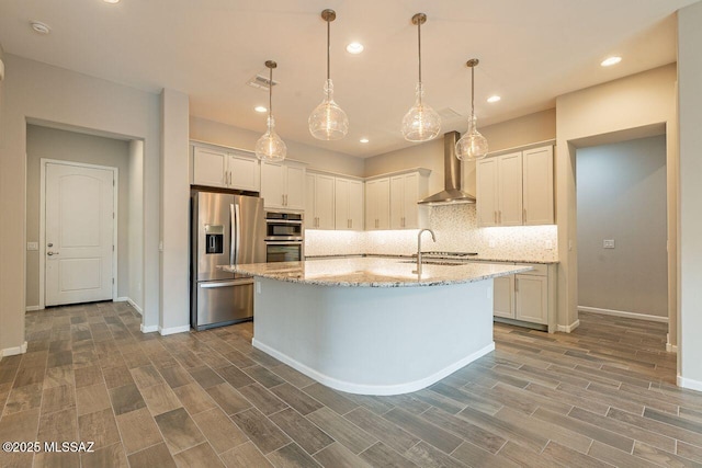 kitchen with a center island with sink, stainless steel appliances, hanging light fixtures, wall chimney exhaust hood, and white cabinets