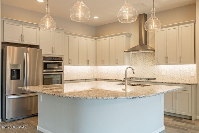 kitchen with stainless steel refrigerator with ice dispenser, wall chimney range hood, a kitchen island with sink, and hanging light fixtures