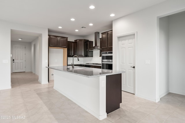 kitchen featuring light stone countertops, dark brown cabinets, stainless steel appliances, wall chimney range hood, and a center island with sink