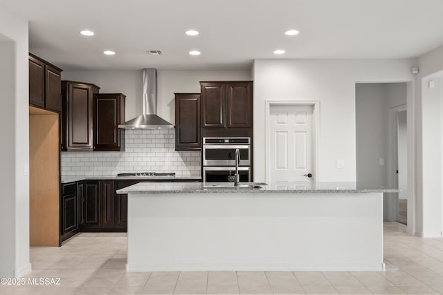 kitchen with backsplash, light stone counters, wall chimney exhaust hood, stainless steel appliances, and a kitchen island with sink