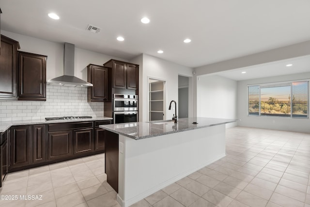 kitchen featuring appliances with stainless steel finishes, wall chimney exhaust hood, sink, an island with sink, and light tile patterned flooring