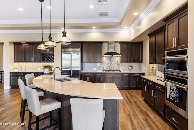 kitchen with a kitchen island with sink, sink, wall chimney exhaust hood, appliances with stainless steel finishes, and decorative light fixtures