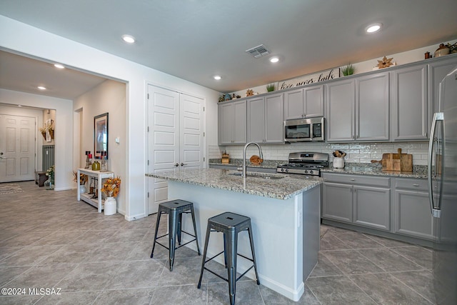 kitchen with gray cabinetry, sink, light stone counters, an island with sink, and appliances with stainless steel finishes