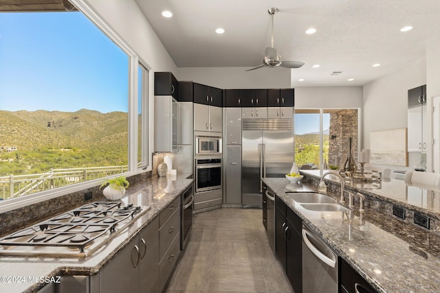 kitchen featuring a mountain view, sink, built in appliances, ceiling fan, and dark stone countertops