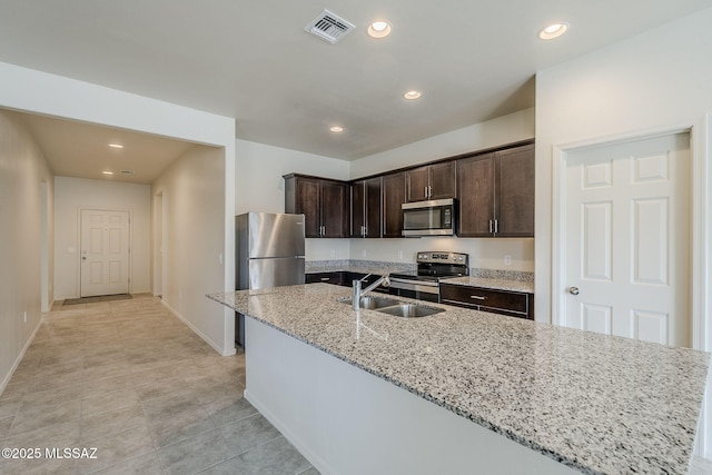 kitchen featuring sink, light stone countertops, an island with sink, appliances with stainless steel finishes, and dark brown cabinets