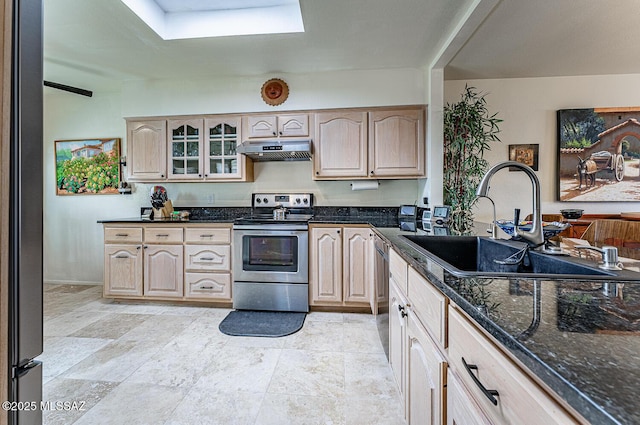 kitchen featuring under cabinet range hood, light brown cabinetry, dark stone countertops, stainless steel appliances, and a sink