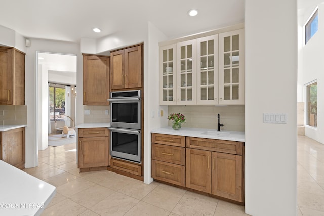 kitchen featuring a sink, double oven, brown cabinetry, light countertops, and glass insert cabinets