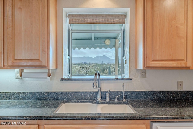 kitchen featuring a textured wall, a sink, stainless steel dishwasher, light brown cabinetry, and dark stone countertops