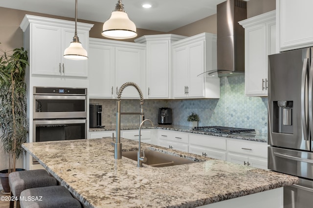 kitchen featuring white cabinets, wall chimney range hood, appliances with stainless steel finishes, tasteful backsplash, and decorative light fixtures