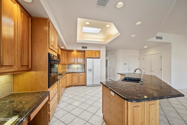 kitchen with a skylight, a center island with sink, black oven, white fridge with ice dispenser, and sink