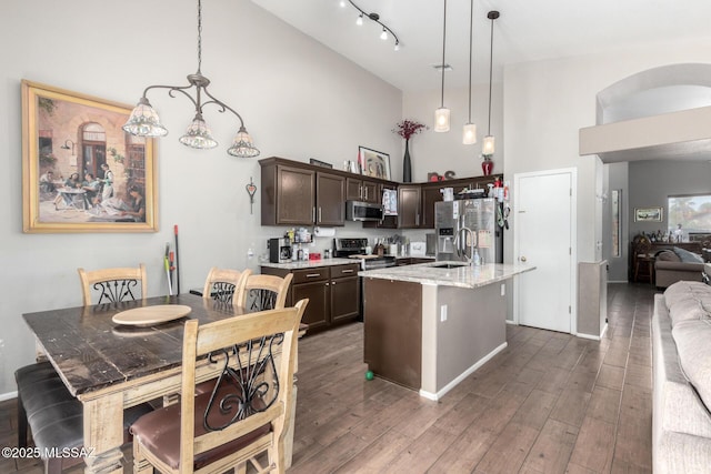 kitchen with appliances with stainless steel finishes, an island with sink, hanging light fixtures, light stone countertops, and dark brown cabinets