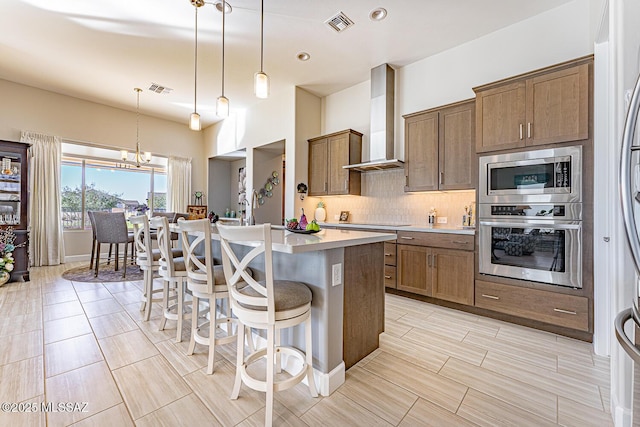 kitchen featuring pendant lighting, wall chimney exhaust hood, stainless steel appliances, an island with sink, and a kitchen breakfast bar