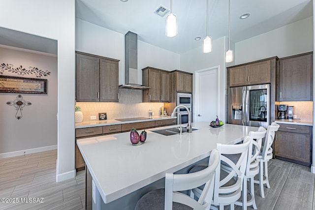 kitchen featuring a kitchen bar, an island with sink, appliances with stainless steel finishes, wall chimney exhaust hood, and sink