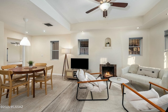 living room with a brick fireplace, a tray ceiling, light hardwood / wood-style floors, and ceiling fan