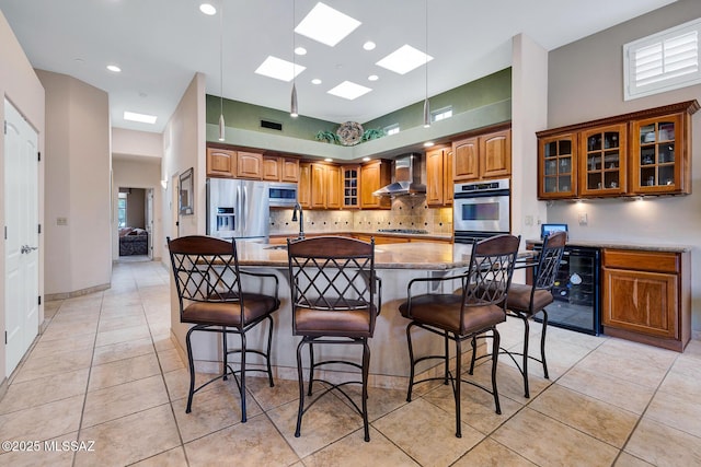 kitchen with an island with sink, wine cooler, stainless steel appliances, light stone countertops, and wall chimney exhaust hood