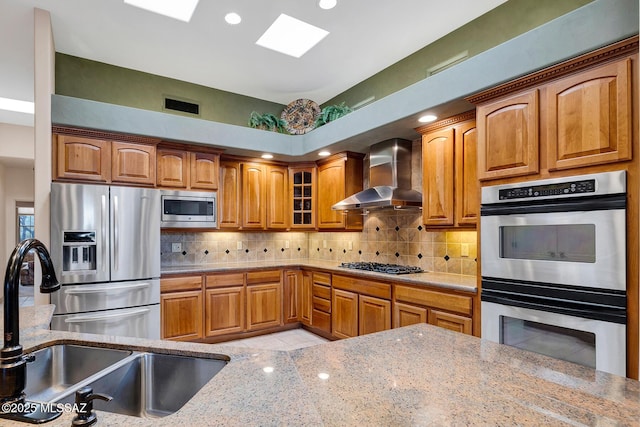 kitchen with wall chimney range hood, sink, appliances with stainless steel finishes, backsplash, and light stone counters