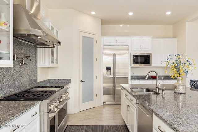 kitchen featuring white cabinetry, wall chimney range hood, light stone counters, built in appliances, and sink