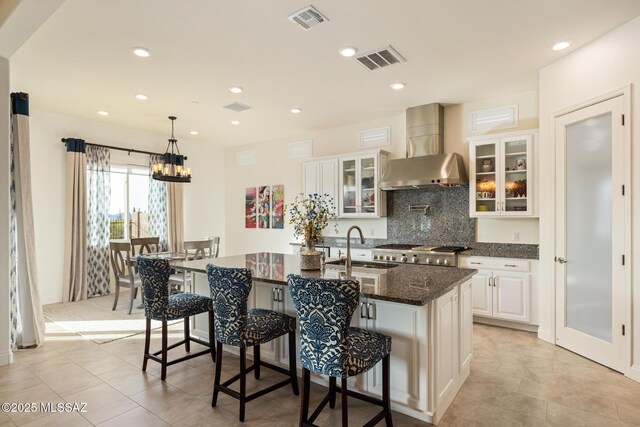 kitchen featuring a kitchen island with sink, dark stone counters, wall chimney range hood, white cabinets, and sink