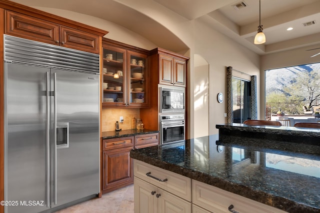 kitchen featuring hanging light fixtures, dark stone countertops, built in appliances, and backsplash