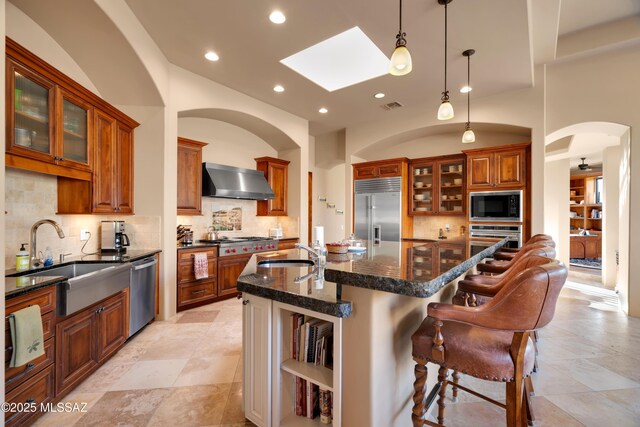 kitchen featuring wall chimney range hood, a skylight, built in appliances, tasteful backsplash, and an island with sink