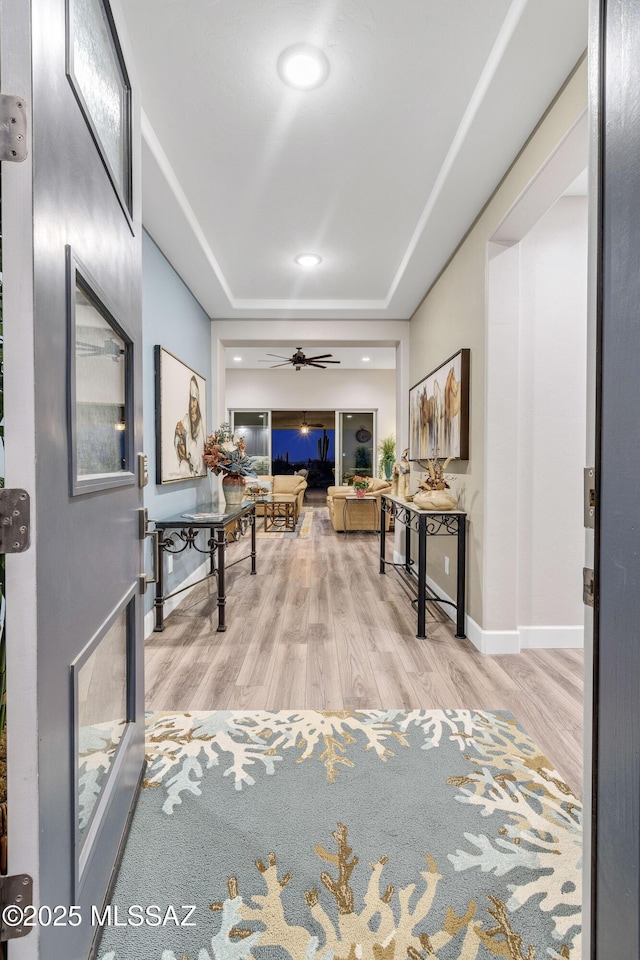 hallway featuring light wood finished floors, recessed lighting, a raised ceiling, and baseboards