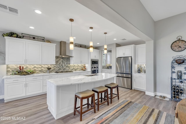 kitchen with visible vents, appliances with stainless steel finishes, white cabinetry, wall chimney range hood, and an island with sink