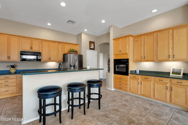 kitchen featuring light brown cabinets, a kitchen island with sink, black appliances, a kitchen breakfast bar, and light tile patterned floors