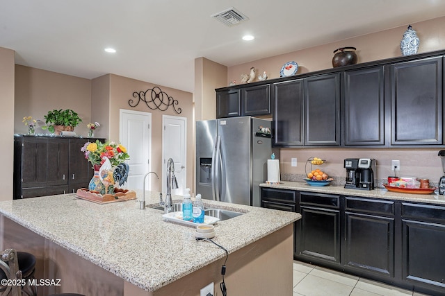 kitchen with a kitchen island with sink, sink, stainless steel fridge, light tile patterned floors, and light stone counters