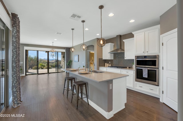 kitchen featuring sink, white cabinets, wall chimney exhaust hood, pendant lighting, and a kitchen island with sink