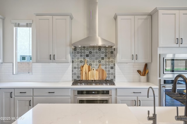 kitchen featuring stainless steel appliances, wall chimney exhaust hood, sink, light stone counters, and decorative backsplash