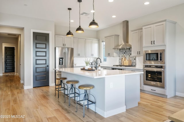 kitchen featuring a center island with sink, decorative light fixtures, wall chimney range hood, stainless steel appliances, and tasteful backsplash